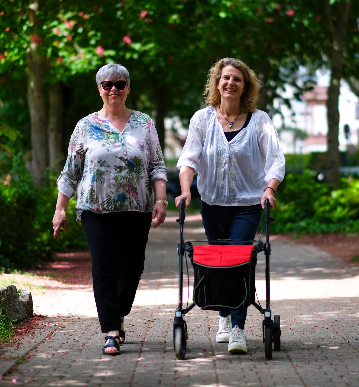 Zwei Frauen beim Spaziergang, eine mit dem Sunrise Gemino 30 Carbon Rollator mit roter City-Tasche (optional)