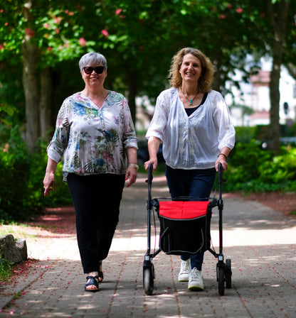 Zwei Frauen beim Spaziergang, eine mit dem Sunrise Gemino 30 Carbon Rollator mit roter City-Tasche (optional)